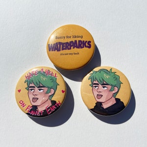 May include: Three round button pins with a yellow background. The first pin has the text "Sorry for liking WATERPARKS it's not my fault". The second pin has a cartoon illustration of a person with green hair and the text "Hate & Hell on Planet Earth". The third pin has a cartoon illustration of a person with green hair.