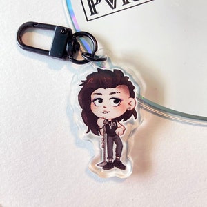 Puede incluir: Un llavero de acrílico transparente con una ilustración de dibujos animados de una persona con cabello castaño, vistiendo una camisa negra y pantalones negros, sosteniendo un micrófono. El llavero tiene un cierre de metal negro.