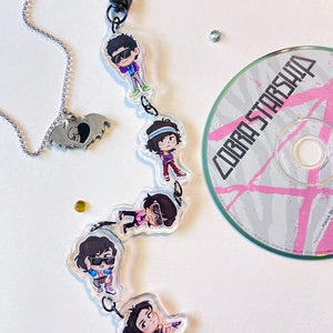 May include: A clear charm bracelet with five cartoon characters wearing sunglasses. The characters are attached to a silver chain with a silver bat charm. The bracelet is displayed on a white surface with a CD labeled "Cobra Starship" in the background.