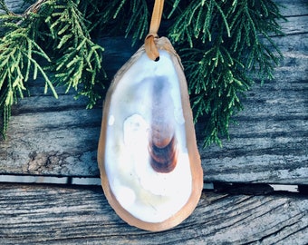 Puede incluir: Adorno artesanal de concha de ostra con interior crema y marrón, bordeado por un borde marrón claro. Se adjunta una cinta dorada para colgar. El adorno se muestra sobre un fondo de ramas verdes de hoja perenne y madera envejecida.