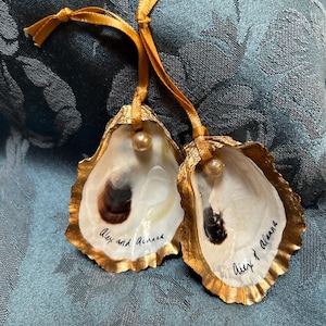 Puede incluir: Dos adornos de concha de ostra con bordes pintados de oro y una pequeña perla dorada. Las conchas tienen un interior nacarado y están inscritas con las palabras "Alex and Alanna" en tinta negra, y están unidas a cintas doradas.