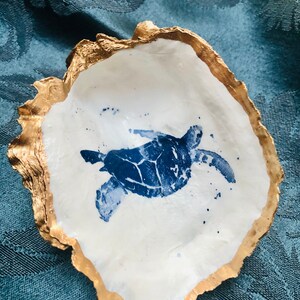 Könnte beinhalten: Eine weiße Keramik-Muschelschüssel mit goldfarbenem Rand und einer blauen Silhouette einer Meeresschildkröte.