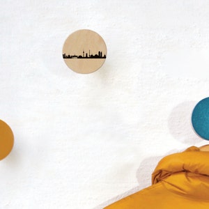 Op de afbeelding: Drie ronde houten wandhaken met een skyline-ontwerp, een is naturel hout, een is geel en een is blauw.