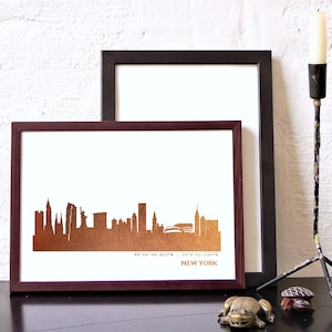 Könnte beinhalten: Ein gerahmter Druck der Skyline von New York City in Kupferfolie. Der Druck ist in einem dunkelbraunen Rahmen und steht auf einem Tisch mit einer kleinen Froschfigur und einer Schildkrötenfigur.