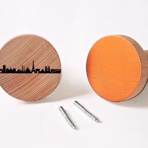 Op de afbeelding: Twee houten wandhaken, een met een zwart silhouet van de skyline van Parijs en de andere oranje geverfd. Beide haken worden geleverd met schroeven voor installatie.