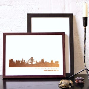 K&ouml;nnte beinhalten: Ein gerahmter Druck der Skyline von San Francisco in Goldfolie auf wei&szlig;em Hintergrund. Der Rahmen ist aus dunkelbraunem Holz.