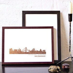 K&ouml;nnte beinhalten: Ein gerahmter Druck der Skyline von San Francisco mit der Golden Gate Bridge in Goldfolie. Der Druck ist in einem dunkelbraunen Rahmen und liegt auf einer schwarzen Oberfl&auml;che.