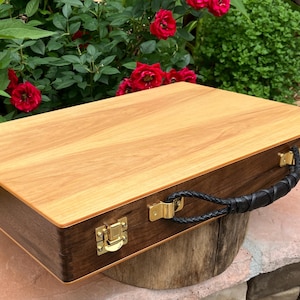 May include: A rectangular wooden carrying case featuring a light-toned wood grain top panel and dark-toned wood sides. The case includes a black braided handle, gold-colored metal latches, and gold-colored metal hinges along the front edge.
