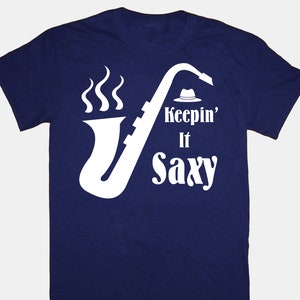 Puede incluir: Una camiseta azul marino con un gráfico de saxofón blanco y el texto "Keepin' It Saxxy".