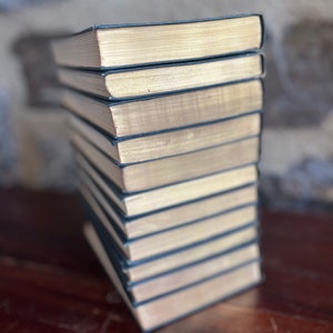 May include: A stack of ten hardback books with black covers and gold-colored edges. The books are arranged vertically on a wooden surface.