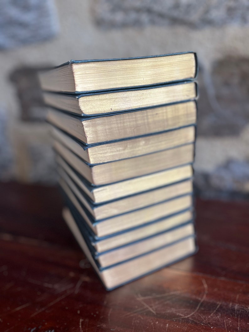 May include: A stack of ten hardback books with black covers and gold-colored edges. The books are arranged vertically on a wooden surface.