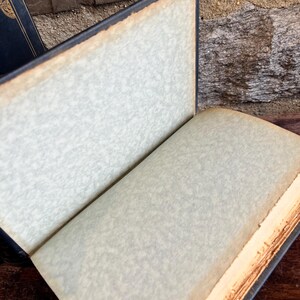 May include: An open antique book with aged, off-white pages and a dark blue cover. The edges of the pages are a warm, golden brown, and the cover shows signs of wear. The book is set against a textured background.