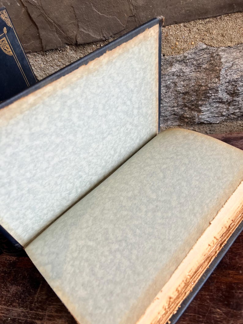 May include: An open antique book with aged, off-white pages and a dark blue cover. The edges of the pages are a warm, golden brown, and the cover shows signs of wear. The book is set against a textured background.
