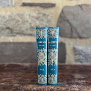 May include: Two vintage cookbooks, titled "America Cook," stand upright. The books have teal covers with gold ornate designs and lettering. The spines display "Vol. I" and "Vol. II."