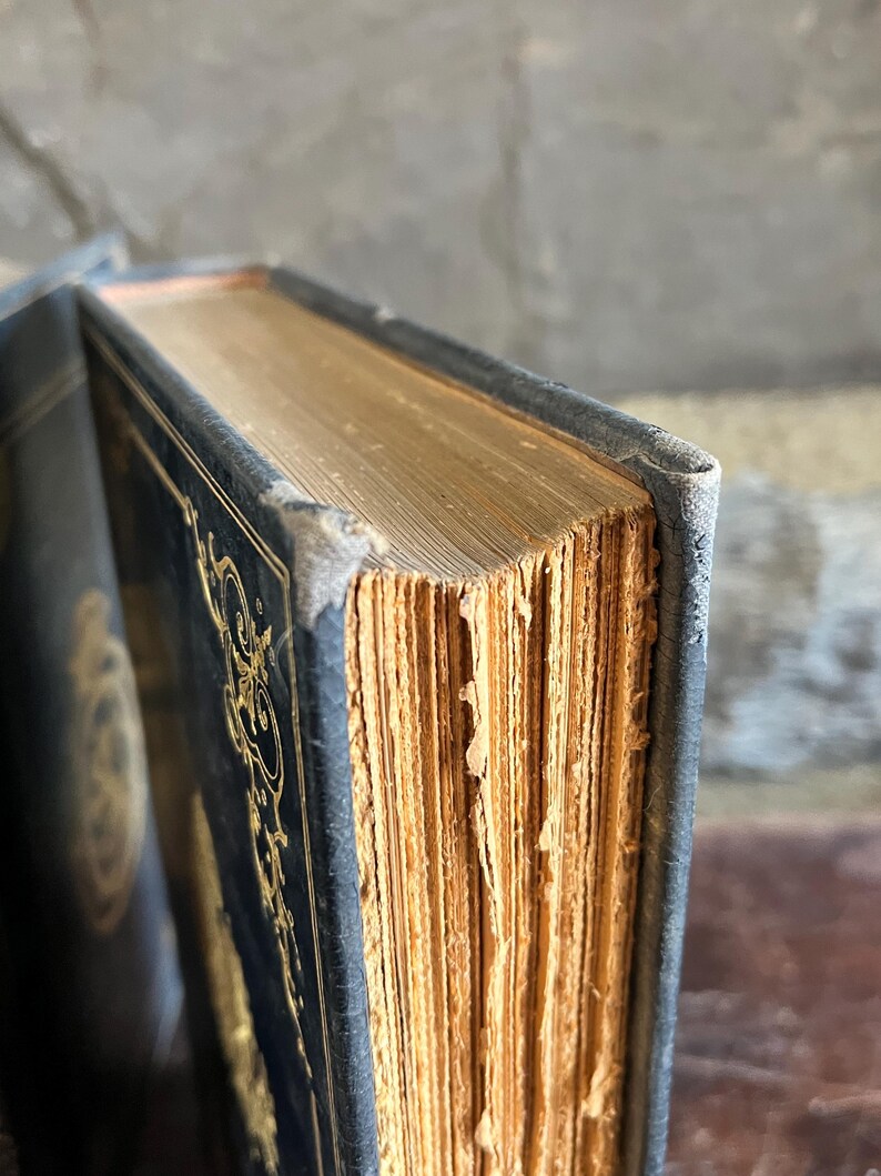 May include: Close-up of an antique book with a dark blue cover and gold embossed designs. The aged pages have a golden hue, and the edges show wear. The book's spine is visible, and the corner is slightly worn.