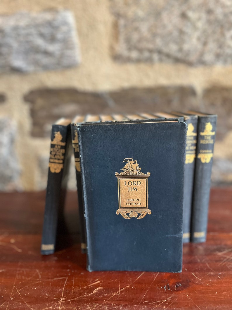 May include: A set of six vintage hardcover books with black covers and gold lettering. The title of the book in the center is "Lord Jim" by Joseph Conrad.