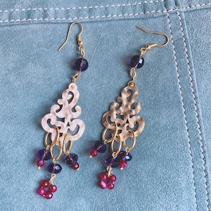 Peut inclure: Une paire de boucles d'oreilles pendantes avec des crochets dorés. Les boucles d'oreilles présentent un motif de filigrane crème, des perles violettes et rouges, et de petits cerceaux dorés. Le fond est un matériau bleu clair semblable à du daim.