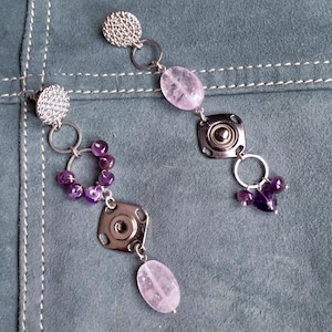 May include: A pair of asymmetrical earrings with silver-tone metal and purple and pink gemstones. Each earring features a textured round stud, connected to a series of rings, beads, and a square metal piece with a central design, and a dangling oval gemstone.
