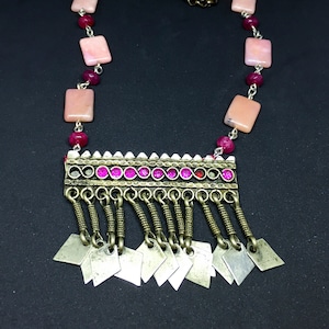 Pode incluir: Um colar rosa e prateado com um pingente retangular que apresenta uma franja de pingentes prateados. O pingente é decorado com contas rosa e prateadas.