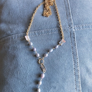 May include: A gold-tone chain necklace featuring a Y-shaped drop design. The necklace is accented with small, oval-shaped lavender-grey pearls and rectangular, translucent, iridescent shell-like beads spaced along the metal chain links.