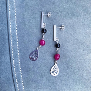 Peut inclure: Une paire de boucles d'oreilles en argent avec fermoir à tige. Chaque boucle d'oreille comporte une perle noire, une perle magenta et un charm en forme de goutte d'eau argenté avec un motif découpé. Les boucles pendent d'une barre droite en argent.