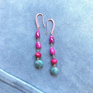 Peut inclure: Une paire de boucles d'oreilles pendantes avec des crochets en or rose et une disposition verticale de trois perles facettées couleur rubis magenta, suivies d'une perle ronde texturée de couleur sarcelle à la base de chaque boucle.