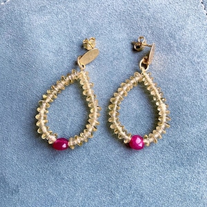 Puede incluir: Un par de pendientes dorados con diseño de lágrima. Los pendientes presentan un borde de piedras preciosas amarillas con cuentas y una sola piedra preciosa rosa redonda en la parte inferior. Se muestran sobre una superficie azul.