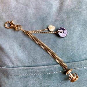 May include: A gold chain keychain with a small gold leaf charm, a black button charm, and a gold anchor charm. The keychain is hanging on a blue fabric surface.