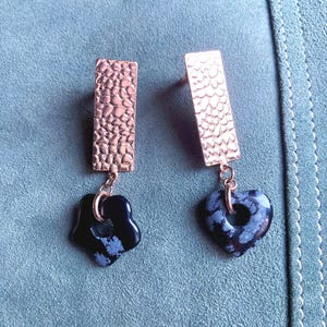 May include: A pair of drop earrings featuring rectangular rose gold-toned metal posts with a textured, hammered surface. Each post suspends a dark, polished stone charm; one is shaped like a five-petaled flower and the other is heart-shaped.