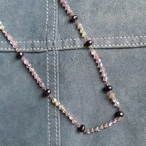 Puede incluir: Un collar delicado con cuentas de cristal iridiscente y perlas negras. El collar está sobre un fondo de ante verde.