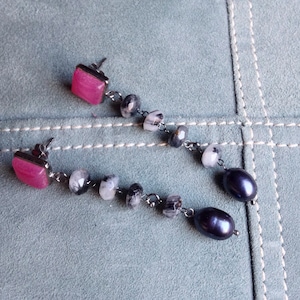 Può includere: Un paio di orecchini pendenti con pietre quadrate rosa e perle nere. Gli orecchini hanno una catena di metallo argentato con perline trasparenti e nere.