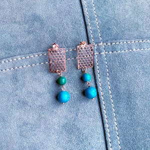 Può includere: Un paio di orecchini color oro rosa con un ciondolo rettangolare in metallo a nido d'ape sospeso a un perno. Ogni orecchino include due perline rotonde, una perlina verde più piccola e una blu più grande, che pendono verticalmente sotto il ciondolo.