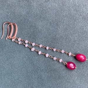 Może przedstawiać: Para kolczyków z różowego złota z delikatnym łańcuszkiem i małymi białymi koralikami. Każdy kolczyk ma różową kropelkę kamienia szlachetnego.