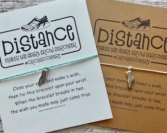 Peut inclure: Une carte blanche avec le texte "Distance makes the heart grow stronger #CROSSCOUNTRY" et un petit charme de chaussure de course argenté attaché à un bracelet en cordon blanc. La carte comprend également des instructions pour faire un vœu avec le bracelet.