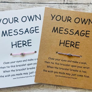 May include: Two cards with the text "Your Own Message Here" printed on them. Each card has a string bracelet attached with a star charm. The card on the left has a pink bracelet and the card on the right has a purple bracelet. The text on the cards instructs the user to close their eyes, make a wish, and tie the bracelet on their wrist. The wish will come true when the bracelet breaks.