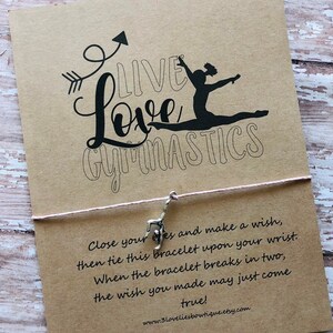 May include: A brown card with the text "Live Love Gymnastics" in black lettering. A silhouette of a gymnast is shown in the middle of the text. The card is tied with a pink string and a silver charm of a gymnast is hanging from the string. The text below the card reads "Close your eyes and make a wish, then tie this bracelet upon your wrist. When the bracelet breaks in two, the wish you made may just come true! www.3loveliesbowtique.etsy.com"
