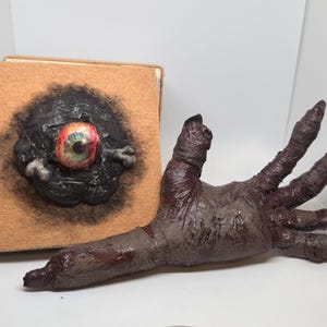 May include: A square tan book with a textured black circular relief on the cover featuring a single realistic eye with a green iris and red veins. Beside the book lies a dark brown, sculpted hand with elongated fingers and a wrinkled, textured surface.