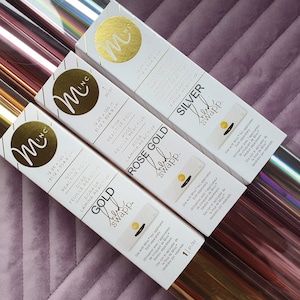 K&ouml;nnte beinhalten: Drei Rollen reaktives Folie zum Basteln in Gold, Ros&eacute;gold und Silber. Jede Rolle ist 31,12 cm x 774,8 cm gro&szlig; und kommt in einer wei&szlig;en Box mit dem Markennamen "Mi-NC" und der Farbe der Folie auf der Vorderseite.