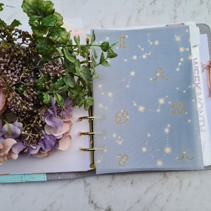 May include: A blue planner with a clear plastic sheet overlay featuring gold zodiac constellations and symbols. The planner has gold rings and a light blue cover. The words "WEEK/MONTH" are visible on the right side of the planner.