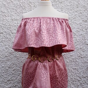 May include: A pink off-the-shoulder top with a leopard print pattern. The top has a ruffled neckline and a gold-colored chain belt. The fabric has a satin sheen. The top is displayed on a mannequin.