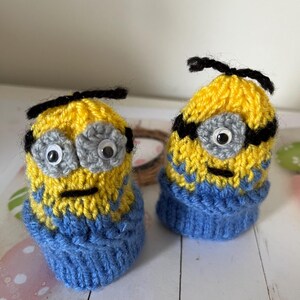 May include: Two knitted Minion characters wearing blue jumpers. The Minions are yellow with black goggles and grey eyes.
