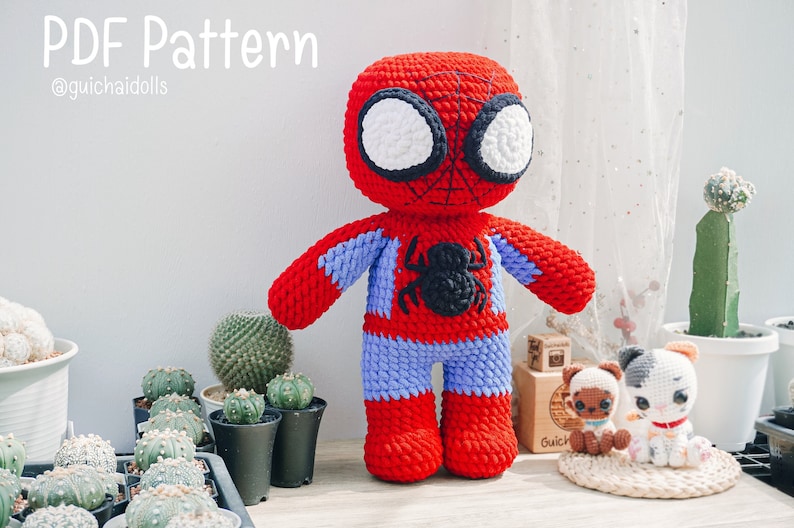 K&ouml;nnte beinhalten: Ein geh&auml;keltes Amigurumi-Spielzeug von Spider-Man in Rot und Blau, mit einem schwarzen Spinnen-Symbol auf der Brust. Das Spielzeug steht auf einer Holzoberfl&auml;che mit einem kleinen Kaktus und anderen Pflanzen im Hintergrund. Der Text "PDF Pattern @guichaiidolls" ist oben im Bild sichtbar.