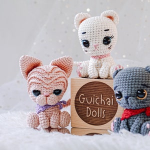 May include: Three crocheted cat toys, one white, one pink, and one gray, sitting on a wooden block with the text "Guichai Dolls".