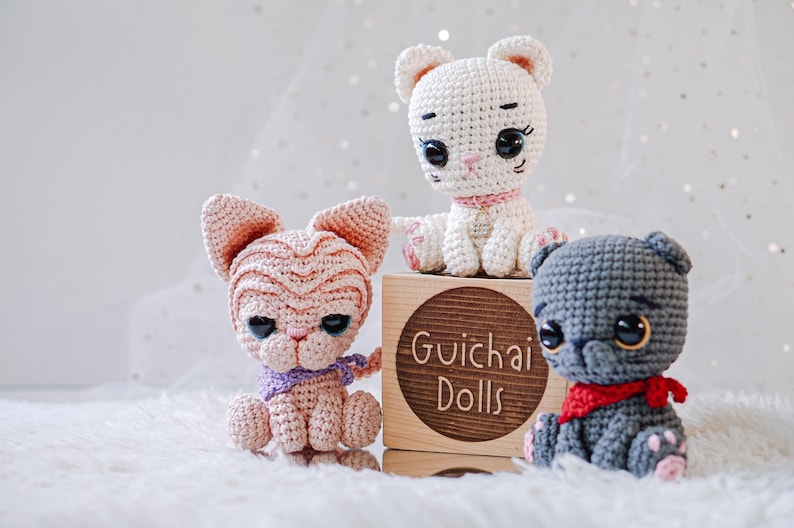 May include: Three crocheted cat toys, one white, one pink, and one gray, sitting on a wooden block with the text "Guichai Dolls".