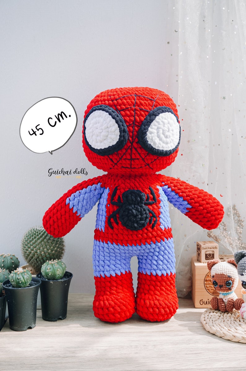 K&ouml;nnte beinhalten: Eine rote und blaue geh&auml;kelte Spiderman-Puppe, 45 cm gro&szlig;, mit einem schwarzen Spinnen-Design auf der Brust. Die Puppe steht auf einer Holzoberfl&auml;che mit einer kleinen Kaktuspflanze in einem schwarzen Topf.