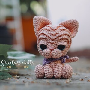 May include: A pink crocheted cat doll with large black eyes and a purple scarf. The doll is sitting on a wooden surface with green leaves in the background. The text "Guichai dolls" is visible in the image.
