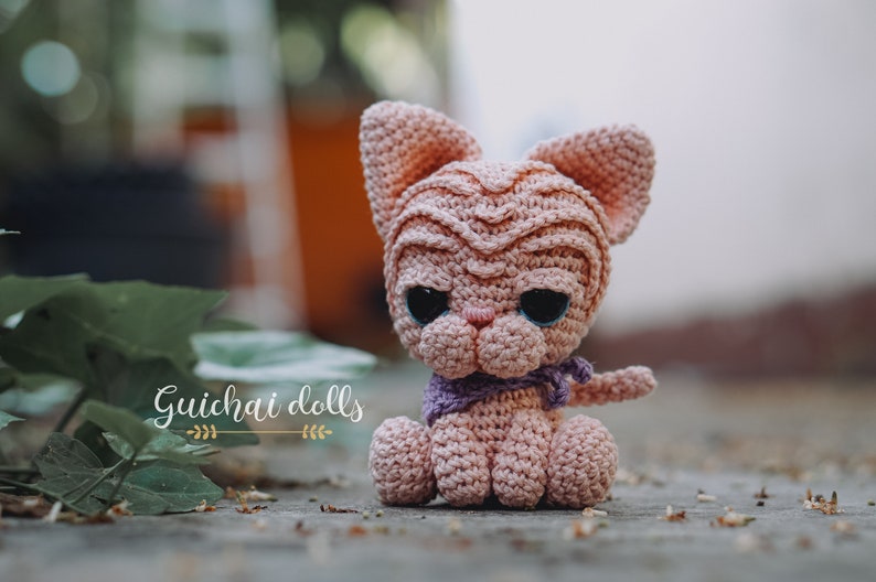 May include: A pink crocheted cat doll with large black eyes and a purple scarf. The doll is sitting on a wooden surface with green leaves in the background. The text "Guichai dolls" is visible in the image.