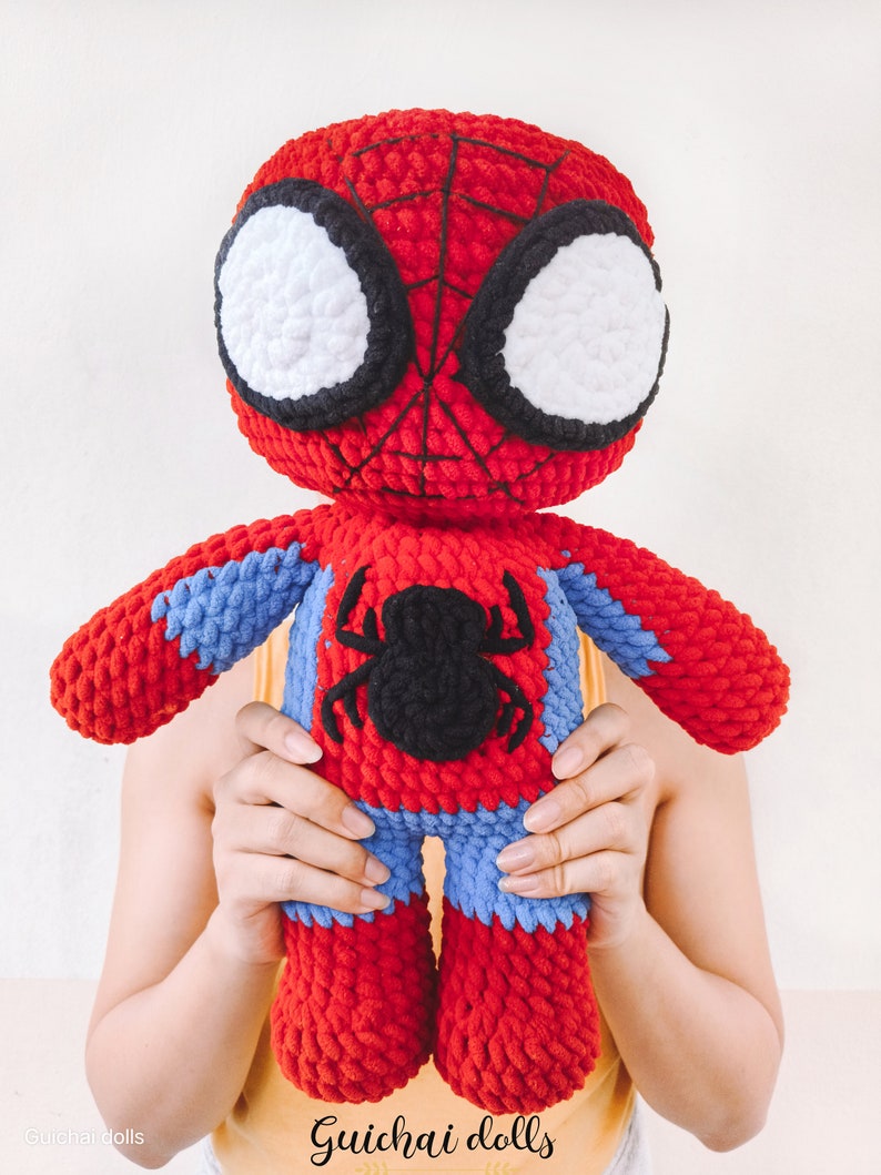 K&ouml;nnte beinhalten: Eine rote und blaue geh&auml;kelte Spiderman-Puppe mit schwarzen Augen und einem schwarzen Spinnenlogo auf der Brust. Die Puppe wird von den H&auml;nden einer Person gehalten. Der Text "Guichai dolls" ist am unteren Rand des Bildes sichtbar.