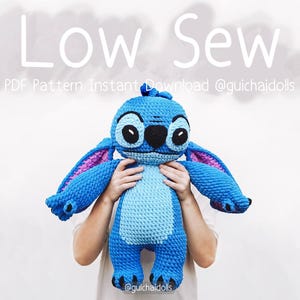 Könnte beinhalten: Eine große, blaue Stitch-Puppe aus Häkelarbeit mit schwarzen Augen und rosa Innenohren wird vor einem weißen Hintergrund gehalten. Die Puppe hat große Ohren und einen strukturierten Körper. Die Worte "Low Sew" stehen oben im Bild.