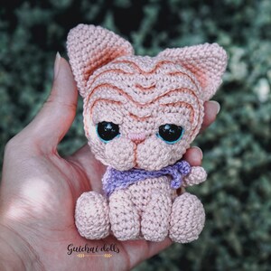 May include: A small crocheted pink cat with blue eyes and a purple scarf. The cat is sitting in a person's hand.